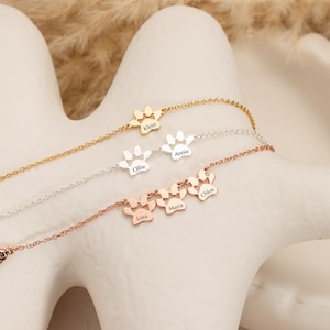 May include: Four delicate necklaces with paw print pendants, each with a different finish: gold, silver, and rose gold. Each paw print has a name engraved: Klein, Ollie, Annie, Lora, Maria, and Chloe. The necklaces are displayed on a white surface.