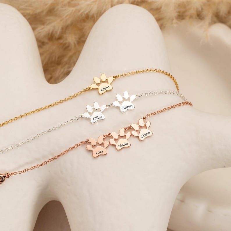 May include: Four delicate necklaces with paw print pendants, each with a different finish: gold, silver, and rose gold. Each paw print has a name engraved: Klein, Ollie, Annie, Lora, Maria, and Chloe. The necklaces are displayed on a white surface.
