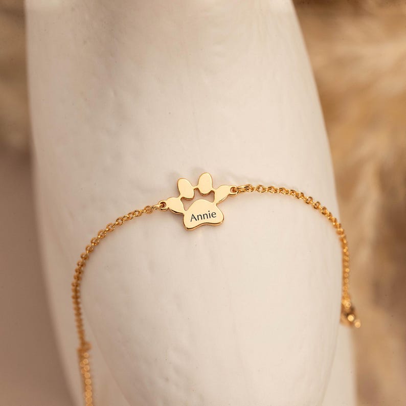 May include: A gold-colored bracelet featuring a paw print charm with the name "Annie" engraved on it. The charm is connected to a delicate chain. The bracelet is displayed against a soft, neutral background.