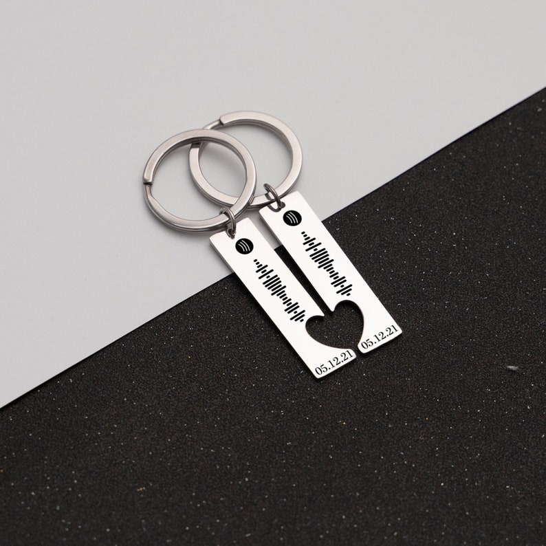 May include: Two silver keychains with a heart-shaped cutout and a bar engraved with a music note and a date. The keychains are connected by a silver ring.