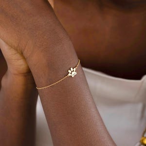 May include: A delicate gold-colored bracelet featuring a paw print charm and a heart-shaped charm with the name "Klein" engraved. The bracelet is worn on a person's arm, showcasing its fine chain and detailed design.
