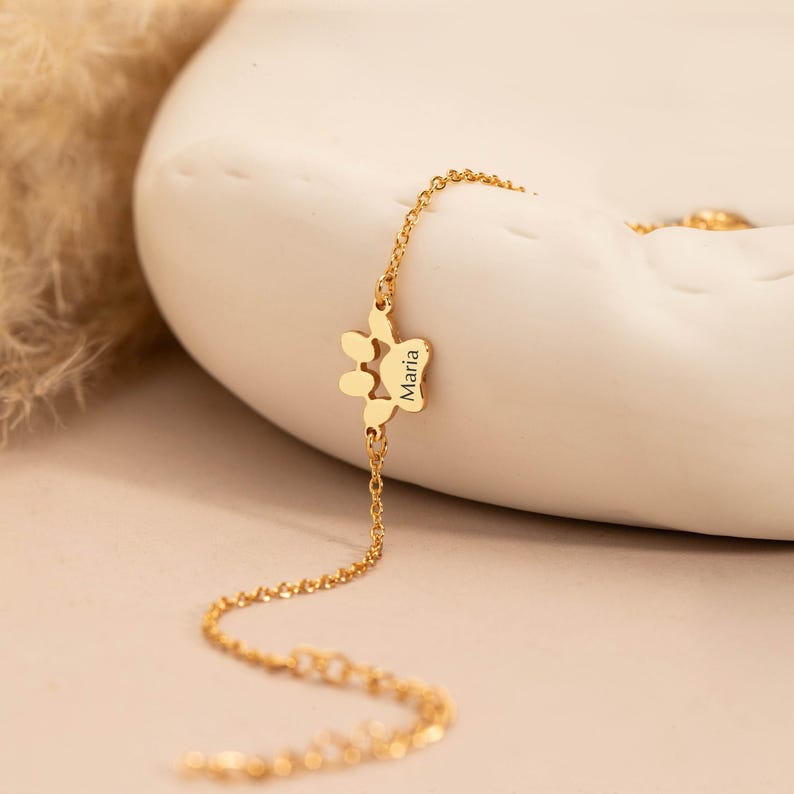May include: A gold-colored bracelet featuring a paw print charm with the name "Maria" engraved on it. The bracelet is displayed on a white, curved surface, with a soft, neutral background. The chain is delicate and the overall aesthetic is elegant.