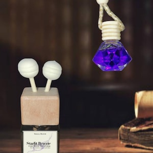 May include: A car reed diffuser with a square wooden top, two white spherical reed sticks, and a label reading Starlit Reverie Car Reed Diffuser. A second diamond-shaped glass bottle filled with purple liquid hangs from a gold-flecked cord.