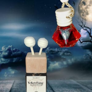 Può includere: Un diffusore per auto con flacone in vetro quadrato, tappo in legno chiaro e due bastoncini bianchi con punta sferica. Un'etichetta riporta la scritta "Small Batch Velvet Fang Car Reed Diffuser". Sopra pende un flacone rosso sfaccettato.
