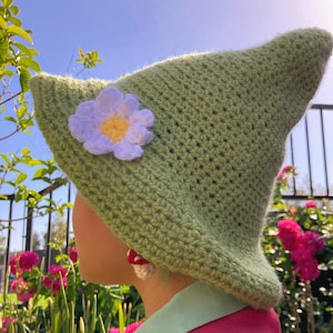 May include: A green crocheted hat with a white flower embellishment. The hat is worn by a person with a red and green shirt.
