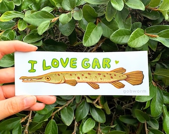 May include: A white sticker with the text "I LOVE GAR" in green letters and a small heart. A cartoon illustration of a brown and yellow gar fish with orange fins is below the text. The artist's name "gobworm" is printed in the bottom right corner.