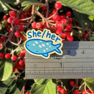 Op de afbeelding: Een houten pin met een blauw en wit gestippeld visontwerp. De pin zegt "She/Her" in gele letters. De pin is ongeveer 2,5 cm lang en wordt gemeten met een liniaal.