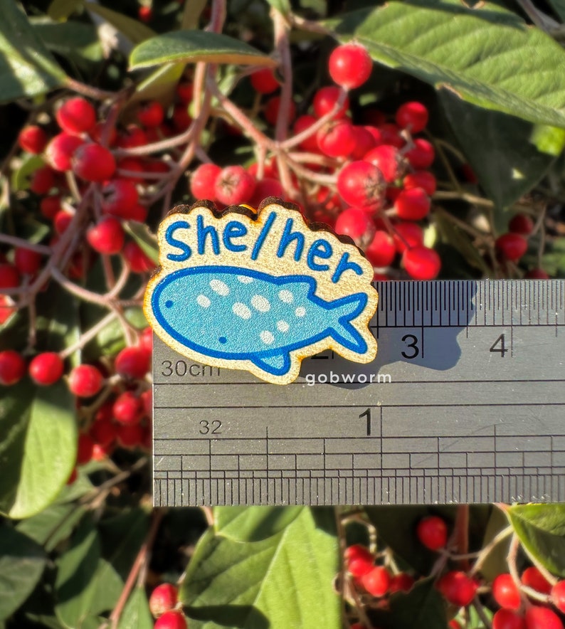 Op de afbeelding: Een houten pin met een blauw en wit gestippeld visontwerp. De pin zegt "She/Her" in gele letters. De pin is ongeveer 2,5 cm lang en wordt gemeten met een liniaal.