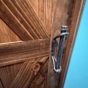 May include: Close-up of a wooden door with a rustic, farmhouse style. The door features a diagonal brace design and a metal handle. The wood has a warm, brown stain, and the metal handle is a dark gray color.
