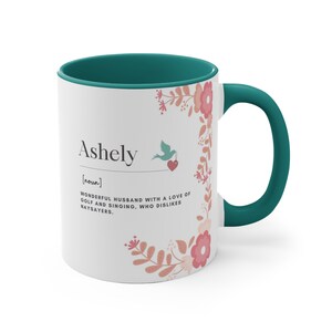 Op de afbeelding: Een witte keramische mok met een teal rand en handvat. De mok heeft een bloemenontwerp en de tekst "Ashely [zelfstandig naamwoord] GEWELDIGE ECHTGENOOT MET EEN LIEFDE VOOR GOLF EN ZINGEN. DIE NIET VAN NEINZEGGERS HOUDT."