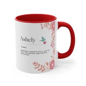 Op de afbeelding: Een witte keramische koffiemok met een rode rand en handvat. De mok heeft een bloemenmotief en de tekst "Ashely [zelfstandig naamwoord] GEWELDIGE ECHTE MAN MET EEN LIEFDE VOOR GOLF EN ZINGEN. DIE NIET VAN NEINZEGGERS HOUDT."