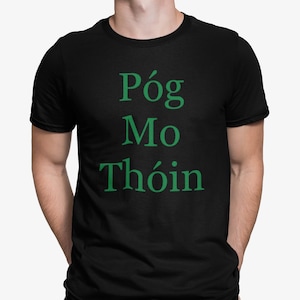 May include: Black t-shirt with the green text "Póg Mo Thóin". The text is stacked vertically. The shirt is a crew neck style and appears to be made of a soft material. The shirt is being worn by a person.