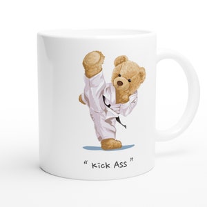 Könnte beinhalten: Weiße Keramiktasse mit einer Cartoon-Illustration eines braunen Teddybären, der einen weißen Karate-Anzug und einen schwarzen Gürtel trägt und mit dem rechten Bein tritt. Der Text "Kick Ass" ist unter dem Bären gedruckt.