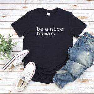 K&ouml;nnte beinhalten: Ein schwarzes T-Shirt mit dem Text "be a nice human." in wei&szlig; auf der Vorderseite. Das Shirt liegt flach auf einer Holzoberfl&auml;che mit einem Paar wei&szlig;en Sneakers und einer blauen Jeans.