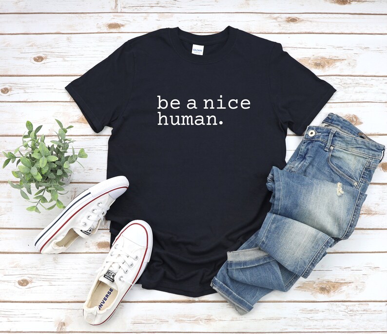 K&ouml;nnte beinhalten: Ein schwarzes T-Shirt mit dem Text "be a nice human." in wei&szlig; auf der Vorderseite. Das Shirt liegt flach auf einer Holzoberfl&auml;che mit einem Paar wei&szlig;en Sneakers und einer blauen Jeans.
