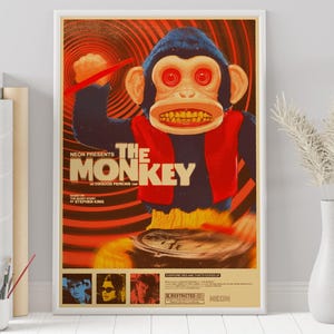 May include: A framed movie poster for "The Monkey" featuring a menacing toy monkey with red eyes and a wide grin. The poster has a retro design with the title in large letters and the text "Neon Presents".
