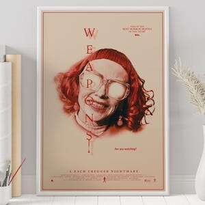 May include: A framed movie poster in red and beige tones. It features a woman with red hair and glasses, with the word "WEAPONS" in red over her face. The text at the bottom reads "A ZACH CREGGER NIGHTMARE."