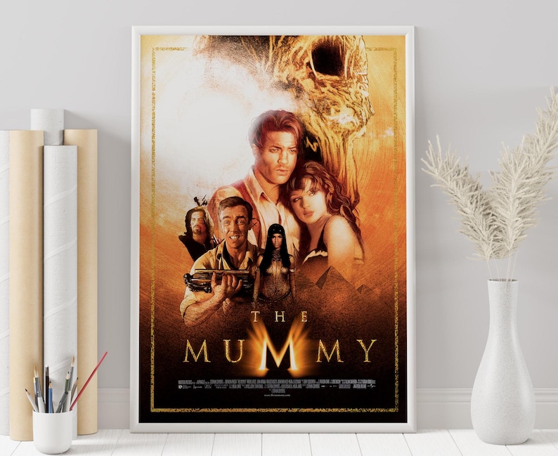 May include: Movie poster for the film "The Mummy" featuring a group of people in a desert setting with a large, golden, skeletal head in the background. The title "The Mummy" is written in large, bold letters at the bottom of the poster.