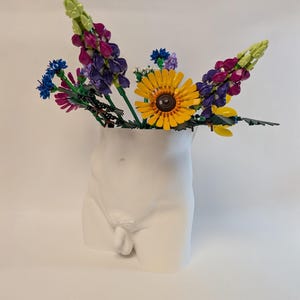Può includere: Un vaso di ceramica bianca a forma di torso maschile, riempito di fiori artificiali colorati. I fiori includono girasoli gialli, lupini viola e rosa e fiordalisi blu. Il vaso è liscio e dettagliato.