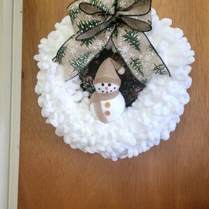 May include: A handmade winter wreath featuring a fluffy white base. A small snowman with a brown hat and scarf is centered. A large bow with a green tree and snowflake pattern tops the wreath, creating a festive holiday decoration.