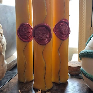 Può includere: Tre alte candele a colonna gialle con accenti argentati e sigilli di cera rossi. Le candele hanno un aspetto rustico e fatto a mano, con un design ondulato. Le candele sono un oggetto decorativo.