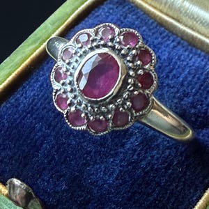 Op de afbeelding: Een zilveren ring met een centrale ovaal geslepen robijn, omringd door tien kleinere rond geslepen robijnen. De zetting heeft een geschulpte rand met kralenachtige metalen details. De ring wordt getoond in een groen doosje met blauwe voering.