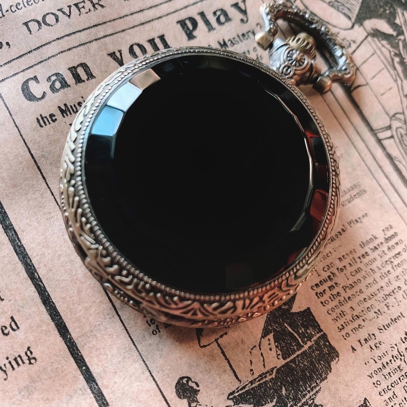 May include: Antique pocket watch with a large, black, faceted face. The watch has an ornate, gold-toned bezel with a decorative pattern. The watch is set against a vintage newspaper background.