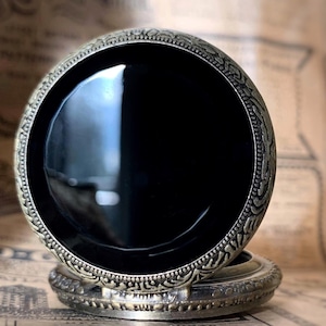 May include: A vintage-style pocket watch with a black face and ornate, antique-gold colored metal casing. The watch has a detailed, engraved design around the bezel and on the base. The watch is closed, showing the polished black face.
