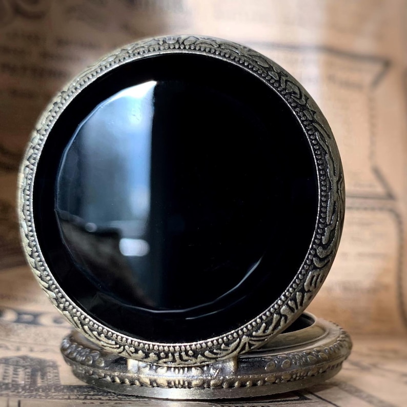 May include: A vintage-style pocket watch with a black face and ornate, antique-gold colored metal casing. The watch has a detailed, engraved design around the bezel and on the base. The watch is closed, showing the polished black face.