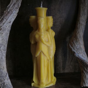 Può includere: Una candela di cera d'api gialla a forma di tre figure in abiti fluenti. La candela ha una parte superiore cilindrica alta con uno stoppino. Le figure sono in cerchio, con le mani giunte. Lo sfondo è in legno scuro.