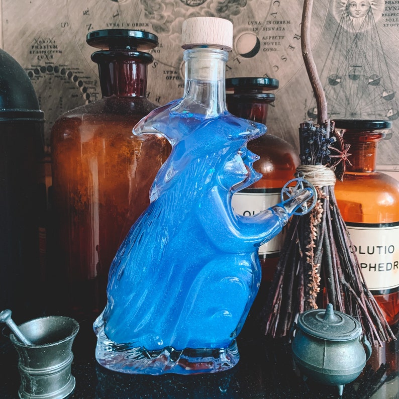 K&ouml;nnte beinhalten: Eine blaue Glasflasche in Form einer Hexe, die ein kleines Rad h&auml;lt. Die Flasche ist umgeben von anderen Flaschen, einem Besen und einem kleinen Kessel.