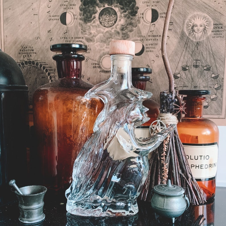 K&ouml;nnte beinhalten: Eine durchsichtige Glasflasche in Form einer Hexe mit einem Korkstopfen. Die Flasche steht auf einem Tisch mit anderen Flaschen und einem Besen. Der Hintergrund ist eine Vintage-Illustration von Mond und Sternen.