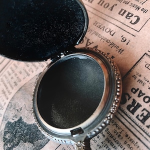 May include: Open silver-toned locket with a black interior and a decorative floral border. The locket is open, revealing the dark interior, and rests on a vintage newspaper background. The locket's design is intricate and detailed.