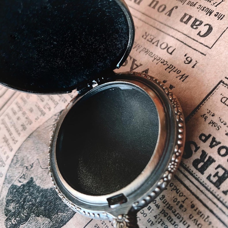 May include: Open silver-toned locket with a black interior and a decorative floral border. The locket is open, revealing the dark interior, and rests on a vintage newspaper background. The locket's design is intricate and detailed.