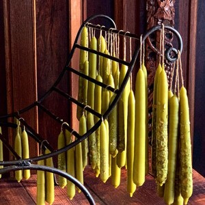 Puede incluir: Un portavelas de metal con un diseño curvo sostiene una colección de velas largas, delgadas y de cera de abeja amarilla. Las velas cuelgan del portavelas y todas son del mismo tamaño.