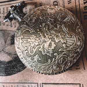 May include: Antique silver-toned pocket watch with intricate floral and scrollwork detailing. The watch has a textured surface and a hinged lid. The top of the watch has a decorative handle. The background is a vintage newspaper.