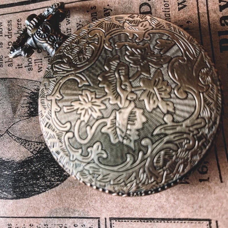 May include: Antique silver-toned pocket watch with intricate floral and scrollwork detailing. The watch has a textured surface and a hinged lid. The top of the watch has a decorative handle. The background is a vintage newspaper.