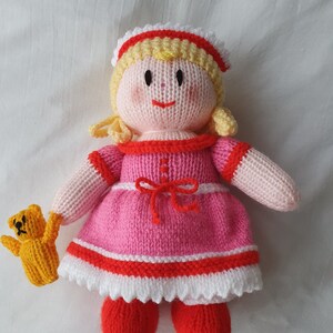 Puede incluir: Una muñeca de punto con pelo rubio, que lleva un vestido rosa con ribetes rojos y blancos, sosteniendo un oso de peluche amarillo de punto.