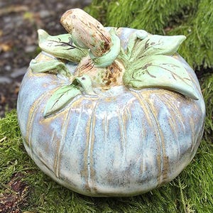 May include: A ceramic pumpkin sculpture featuring a glossy, variegated glaze in shades of pale blue, cream, and earthy brown. The pumpkin has a textured, sculpted stem and several attached green leaves resting on top of its rounded, ribbed body.