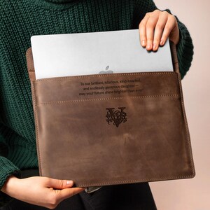 May include: A brown leather laptop sleeve with a stitched border. The sleeve has a quote engraved on it, along with a decorative emblem. The laptop is partially visible inside the sleeve.