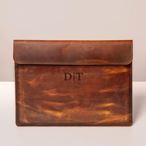 May include: A brown leather laptop sleeve with a flap closure. The sleeve has a distressed finish and features the initials "DT" embossed on the front. The leather has a rich, warm tone.