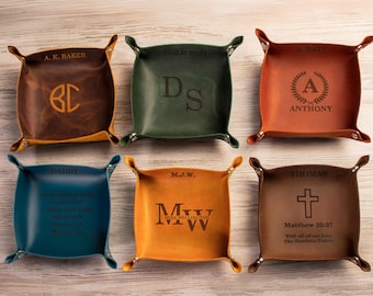 May include: Six square leather valet trays in various colors including dark brown, green, tan, blue, yellow, and medium brown. Each tray features metal snaps at the corners and is personalized with engraved names, initials, dates, or religious text.