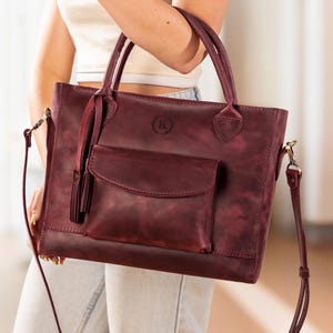 Op de afbeelding: Een bordeauxrode leren tote bag met een voorvak en een afneembare schouderriem. De tas heeft twee handvatten aan de bovenkant en een decoratieve kwast. Een cirkelvormig embleem met de letter "K" is zichtbaar op de voorkant van de tas.