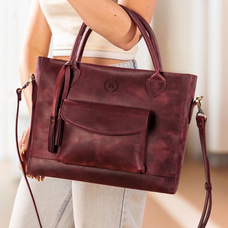 Op de afbeelding: Een bordeauxrode leren tote bag met een voorvak en een afneembare schouderriem. De tas heeft twee handvatten aan de bovenkant en een decoratieve kwast. Een cirkelvormig embleem met de letter "K" is zichtbaar op de voorkant van de tas.