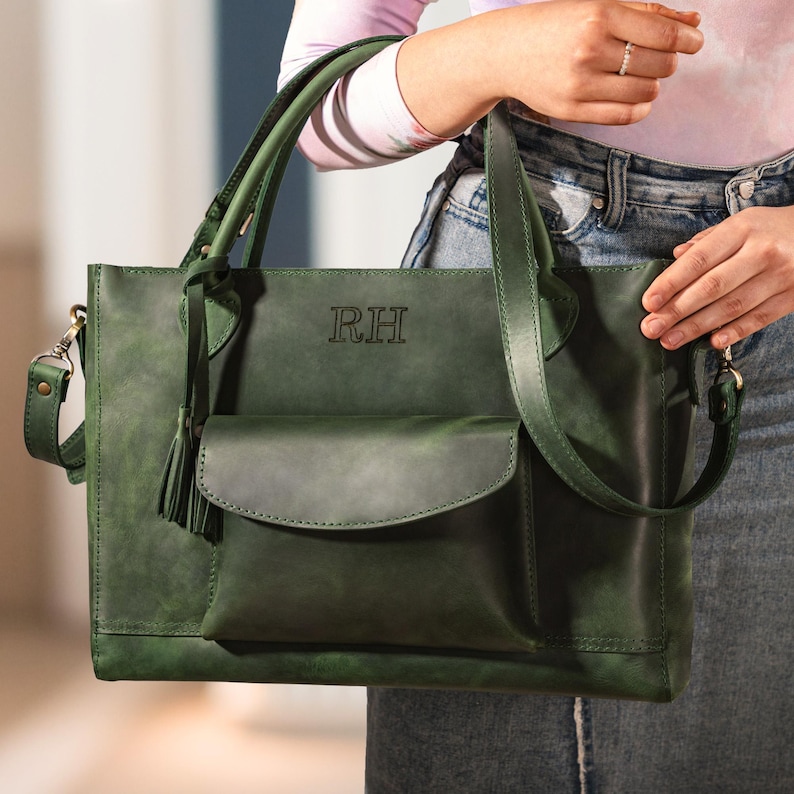 Op de afbeelding: Een groene leren tote bag met een schouderriem en handvatten. De tas heeft een voorvak, een kwast en de initialen "RH" in reli&euml;f op de voorkant. De tas is ongeveer 35 cm breed en 30 cm hoog.