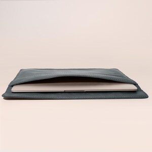 May include: A slate-blue leather laptop sleeve with a stitched border. A silver laptop is partially visible inside the open sleeve. The sleeve is rectangular and lies flat on a light-colored surface.