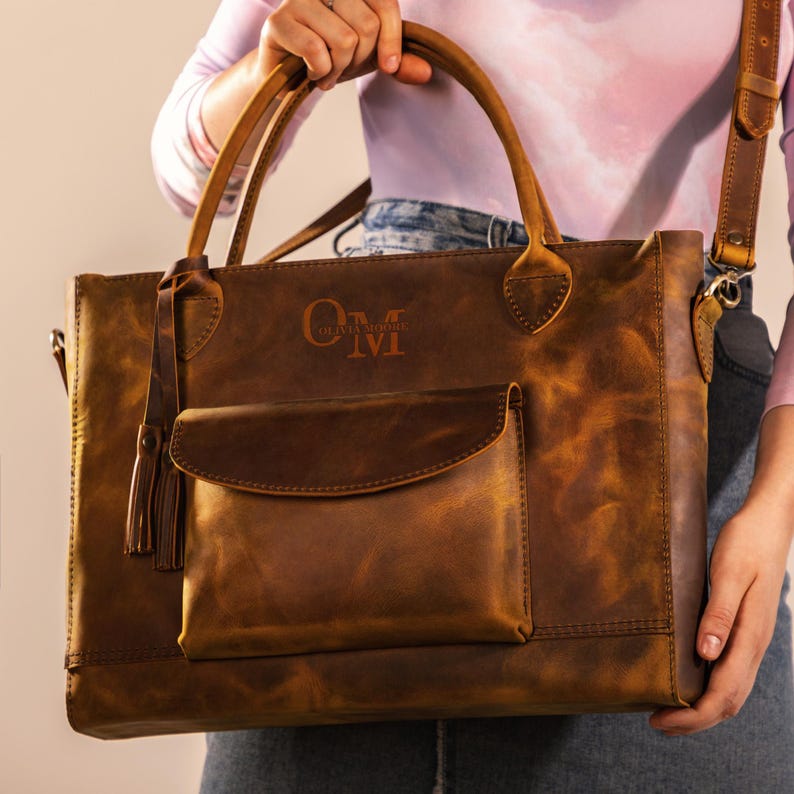 Op de afbeelding: Een bruine leren tote bag met een voorvak en een kwastje. De tas heeft twee handvatten en een schouderriem. De initialen "OM" en "TAT" zijn op de voorkant van de tas in reli&euml;f aangebracht.