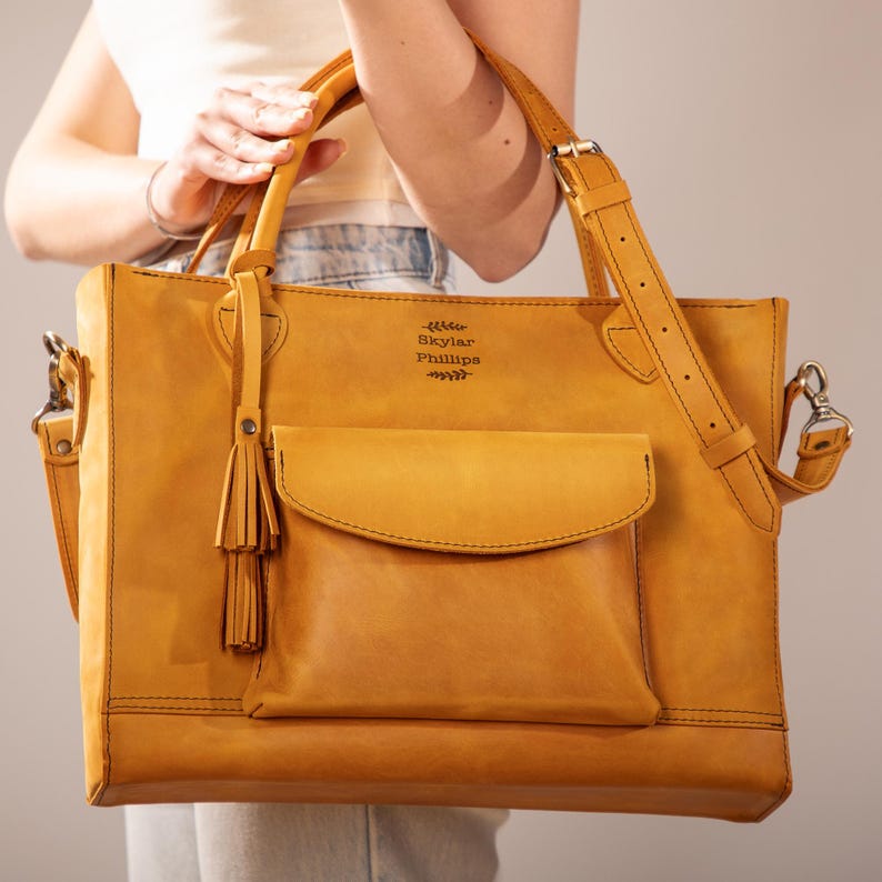 Op de afbeelding: Een mosterdgele leren tote bag met een voorvak en een afneembare schouderriem. De tas heeft een handvat aan de bovenkant en een decoratieve kwast. De naam "Skylar Phillips" is op de bovenkant van de tas in reli&euml;f aangebracht.