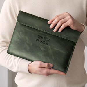 May include: A dark green leather laptop sleeve with a flap closure. The sleeve has the initials "RH" embossed on the front. The leather has a distressed look. The sleeve is held by a person wearing a cream-colored sweater.