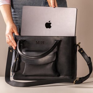 Op de afbeelding: Zwarte leren tote bag met een voorvak, handvatten aan de bovenkant en een afneembare schouderriem. De tas heeft de initialen "M|H" in reli&euml;f op de voorkant en bevat een zilveren laptop. Een zwarte kwast hangt aan de handgreep.